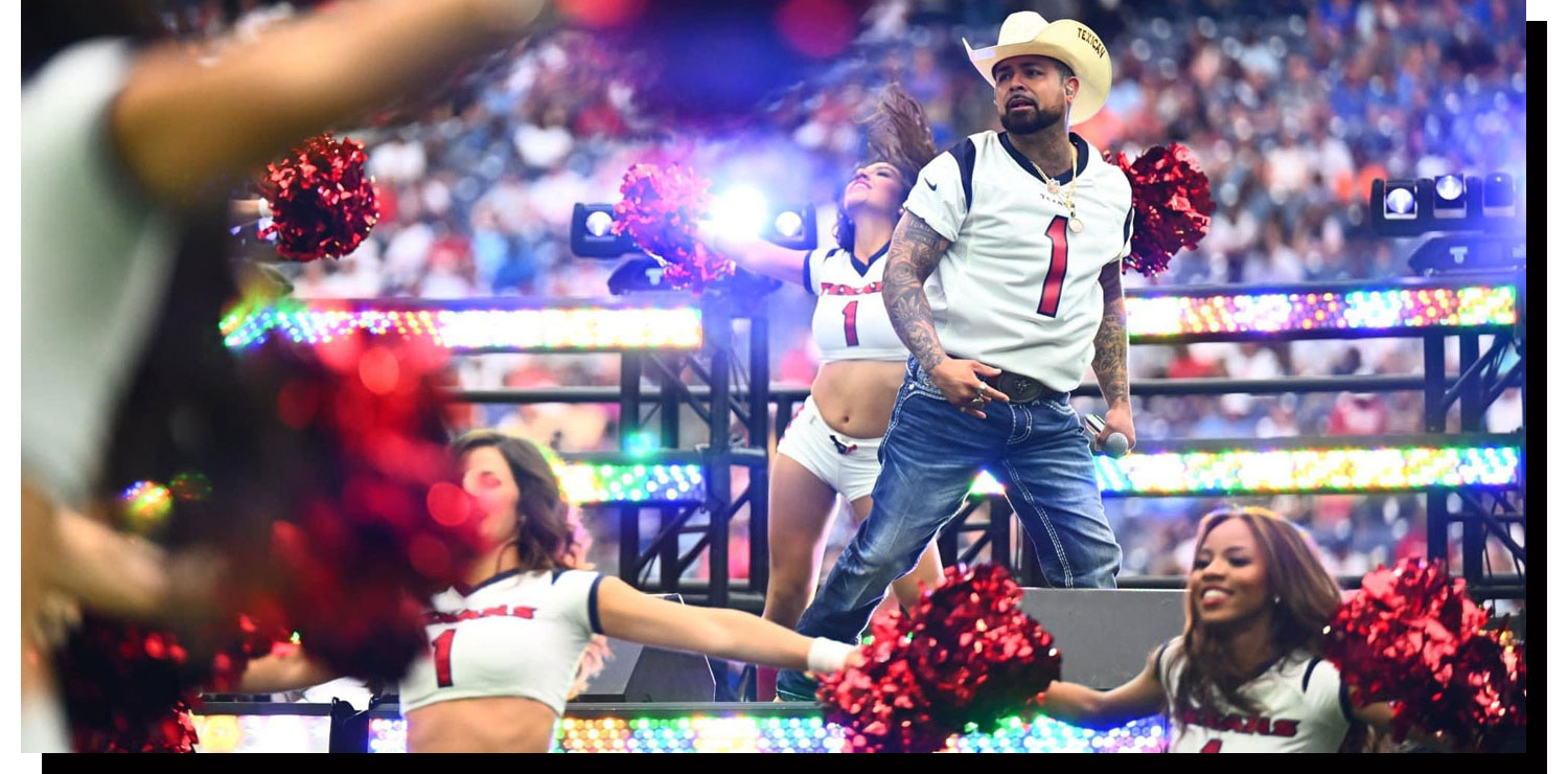 A man, Louie The Singer, dressed as a cowboy, with a straw hat on stage next to dancers/ cheerleaders holding a microphone.